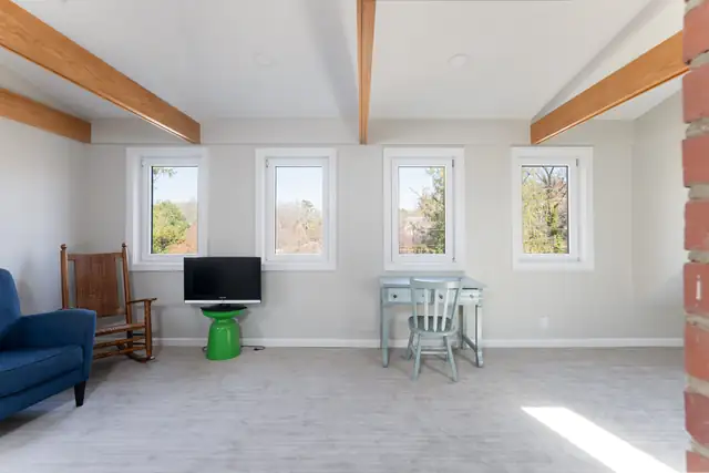 Bright sunroom addition with exposed wood beams, three windows, gray floor and a rocking chair by a brick wall