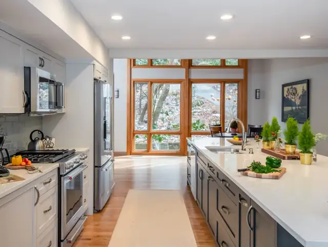 Galley kitchen with white upper cabinets, a gray island with a white quartz top and a wall of wood-framed windows