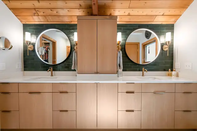 Double vanity in light wood with round mirrors, green tile, brass sconces and a tongue and groove wood ceiling