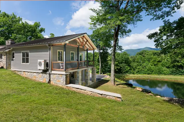 Custom built home addition with a covered porch and stone foundation beside a pond, mountains behind